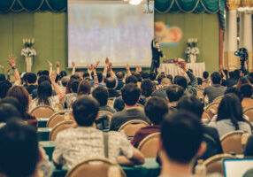 Rear view of Audience showing hand to answer the question from Speaker on the stage in the conference hall or seminar meeting, business and education concept